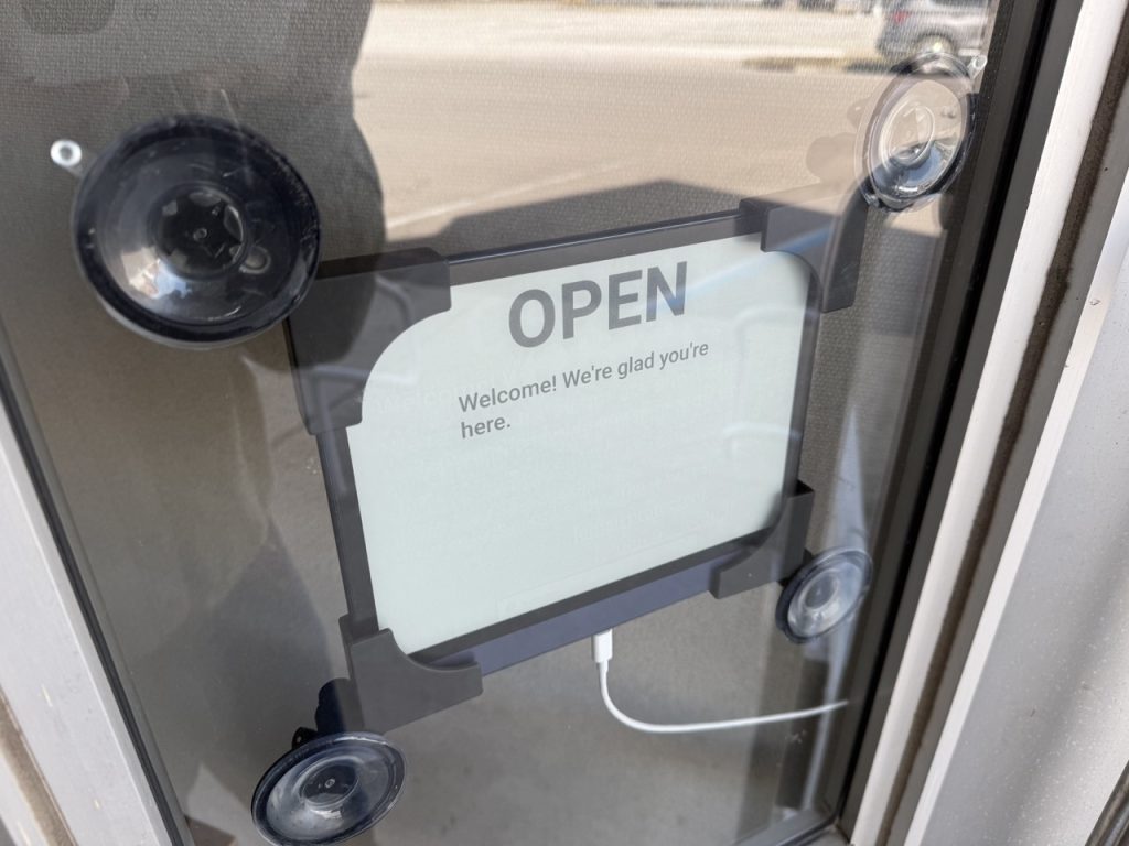 A photo of a BOOX Note Air2 Series mounted in a window as a sign, with the text "OPEN: Welcome! We're glad you're here." on the screen.